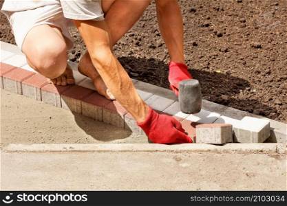 A young worker lays paving slabs on a bright sunny summer day on a prepared sand base on the sidewalk. Copy space.. A worker puts paving slabs on the sidewalk on a bright sunny day.