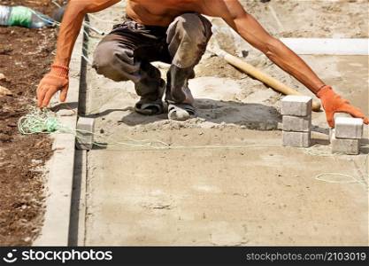 A young worker with a naked, tanned torso pulls nylon thread to lay paving slabs on a bright sunny summer day. Copy space.. A young bare-chested worker uses nylon string to level paving slabs on a bright sunny day.