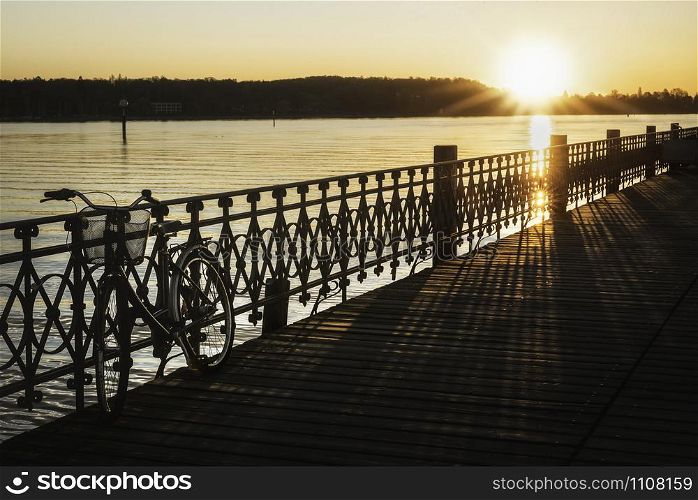Abandoned bicycle on a wooden pier over lake Constance water at sunrise, in Konstanz, Germany. Deck over Bodensee lake at dawn. Sunny day at lake.