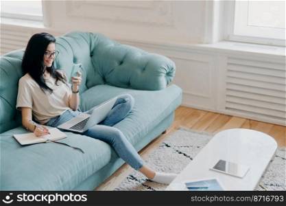 Above shot of pretty brunette woman focused in samrtphone display, writes down necessary information in notebook, watches training webinar, poses in cozy room on comfortable couch, works from home
