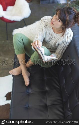 Above view of a beautiful young smiling woman sitting on leather couch relaxing while holding a book at home. Isabella Antonelli