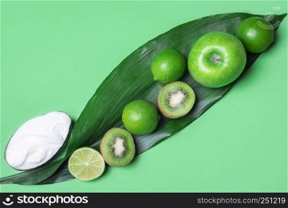 Above view of a green table with a mix of green fruits, apple, kiwi, limes on a big leaf and yogurt. Minimalist style. Healthy eating. Vegan food.