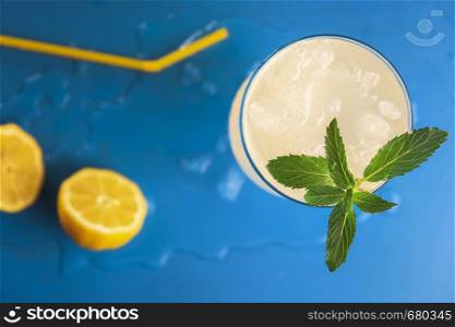 Above view of a lemonade glass with crushed ice and peppermint branch and yellow straw and lemons, on a blue background. Refreshing cold summer drink.