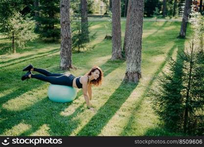 Above view of determined young brunette European woman exercises pilates in green park, balances on fitness ball, dressed in activewear, looks happily somewhere poses outside. Selective focus
