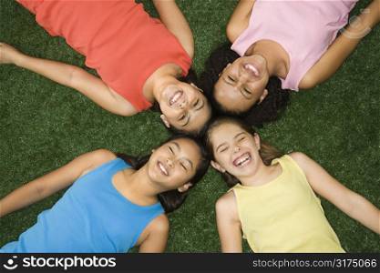 Above view of four girls lying on artificial grass with heads together laughing.
