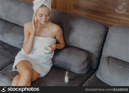Above view of relaxed beautiful young woman sits on comfortable sofa, applies cream on her face, takes care of body, uses cosmetic product. Skin care daily routine. Beauty procedures at home