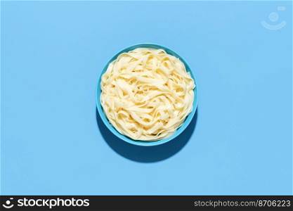 Above view with a bowl full of tagliatelle pasta, minimalist on a blue table. Boiled pasta without sauce in bright light on a colorful background