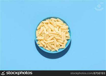 Above view with a penne pasta bowl minimalist on a blue table. Plain cooked pasta, without any sauce, in a blue bowl.
