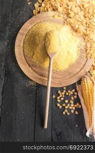Above view with a pile of corn flour on a wooden spoon and trencher, surrounded by corn flakes, grains and corn cob, on a black rustic table.