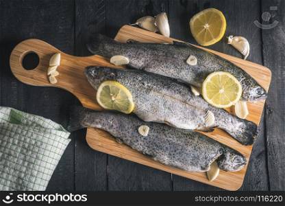 Above view with fresh fish seasoned with garlic and slices of lemons, displayed on a wooden cutting board, on a rustic black table.