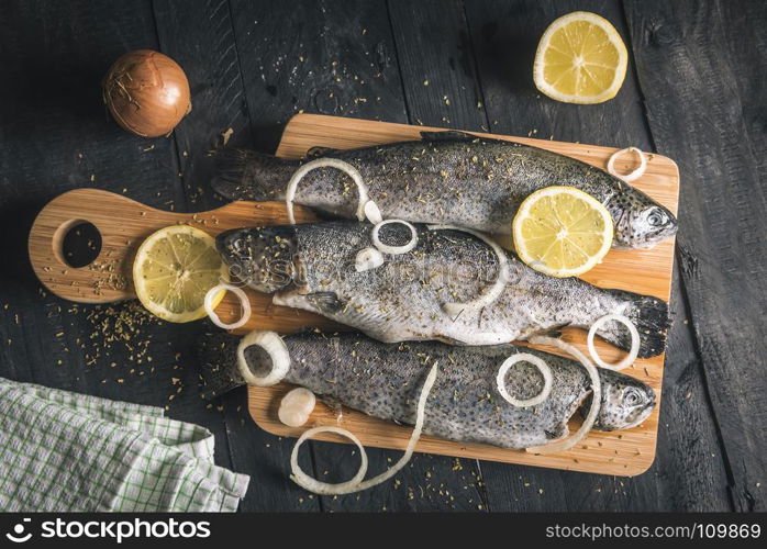 Above view with fresh fish seasoned with spices, herbs, lemon slices and onion rings, on a wooden platter, on a black table, in natural light.