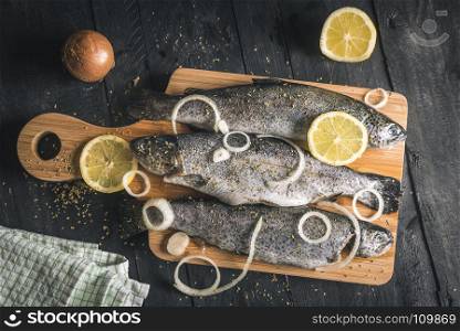 Above view with fresh fish seasoned with spices, herbs, lemon slices and onion rings, on a wooden platter, on a black table, in natural light.