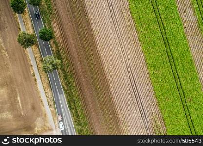 Abstract vertical aerial view of a narrow country road at the edge of an area of arable land with different coloured stripes.