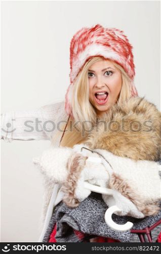 Accessories and clothing for cold days, fashion concept. Blonde woman in winter warm furry hat russian style holding big pile of clothes. Outfit picking problem. Woman in winter hat holding clothes pile
