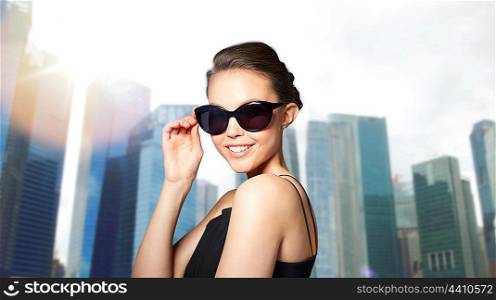 accessories, eyewear, fashion, people and luxury concept - beautiful young woman in elegant black sunglasses over city skyscrapers background