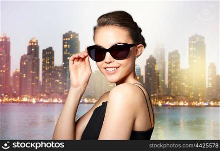 accessories, eyewear, fashion, people and luxury concept - beautiful young woman in elegant black sunglasses over dubai night city lights street background