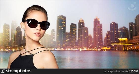 accessories, eyewear, fashion, people and luxury concept - beautiful young woman in elegant black sunglasses over dubai night city lights street background
