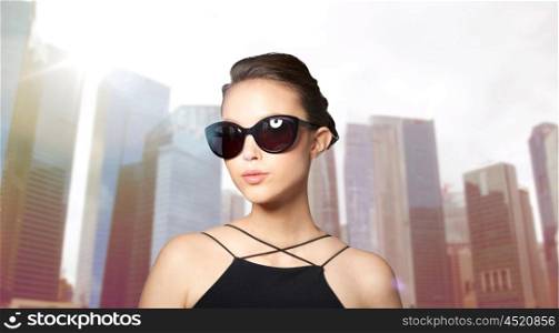 accessories, eyewear, fashion, people and luxury concept - beautiful young woman in elegant black sunglasses over city skyscrapers background