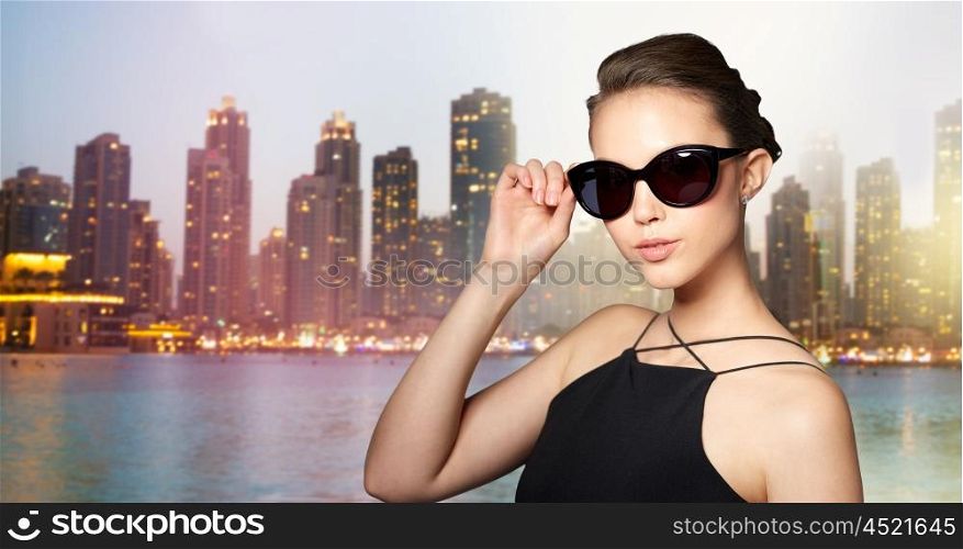 accessories, eyewear, fashion, people and luxury concept - beautiful young woman in elegant black sunglasses over night dubai city lights background