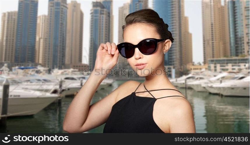 accessories, eyewear, fashion, people and luxury concept - beautiful young woman in elegant black sunglasses over dubai city port background