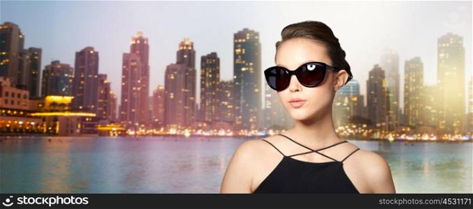 accessories, eyewear, fashion, people and luxury concept - beautiful young woman in elegant black sunglasses over dubai city night lights background