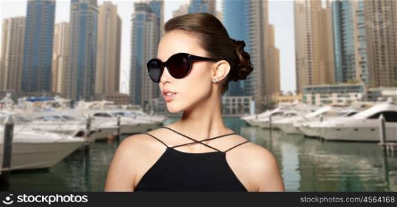 accessories, eyewear, fashion, people and luxury concept - beautiful young woman in elegant black sunglasses over dubai city port background