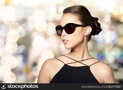 accessories, eyewear, fashion, people and luxury concept - beautiful young woman in elegant black sunglasses over holidays lights background