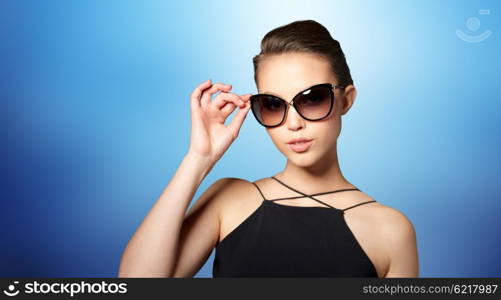 accessories, eyewear, fashion, people and luxury concept - beautiful young woman in elegant black sunglasses over blue background