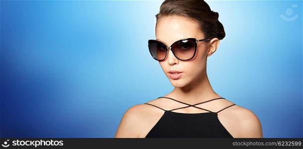 accessories, eyewear, fashion, people and luxury concept - beautiful young woman in elegant black sunglasses over blue background