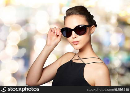 accessories, eyewear, fashion, people and luxury concept - beautiful young woman in elegant black sunglasses over holidays lights background