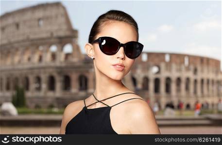 accessories, eyewear, fashion, people and luxury concept - beautiful young woman in elegant black sunglasses over coliseum background