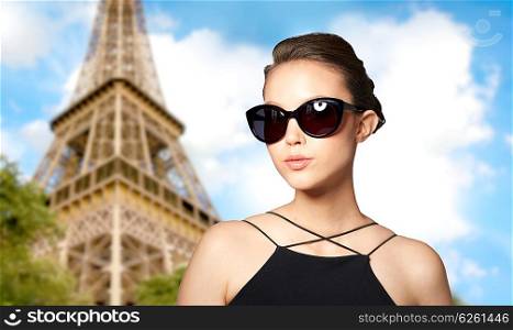 accessories, eyewear, fashion, people and luxury concept - beautiful young woman in elegant black sunglasses over paris eiffel tower background