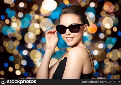 accessories, fashion, people and luxury concept - beautiful smiling young woman in elegant black sunglasses over holidays lights background. beautiful young woman in elegant black sunglasses