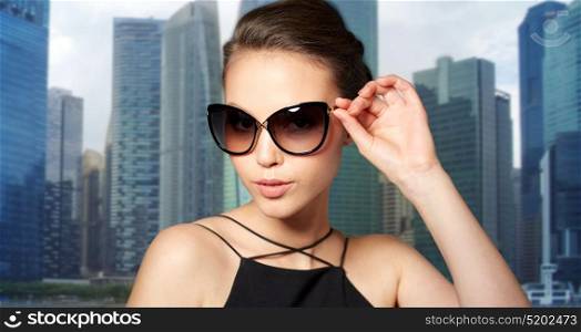 accessories, fashion, people and luxury concept - beautiful young woman in elegant black sunglasses over singapore city skyscrapers background. beautiful young woman in elegant black sunglasses