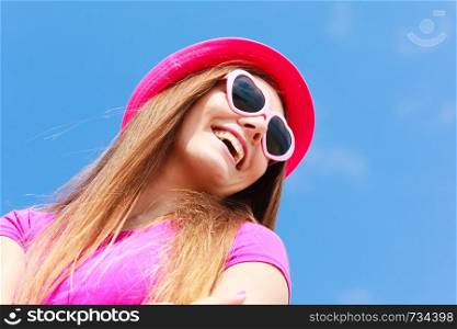 Accessories, female summer fashion, sun protection concept. Smiling beautiful woman wearing heart shaped sunglasses and red hat looking at sky, outdoor shot on sunny day.. Woman wearing heart shaped sunglasses and hat