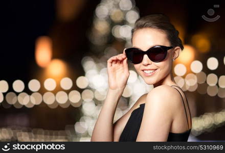 accessories, people and luxury concept - beautiful smiling young woman in elegant black sunglasses over christmas tree lights background. beautiful woman in black sunglasses at christmas