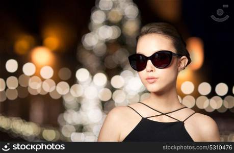 accessories, people and luxury concept - beautiful young woman in elegant black sunglasses over christmas tree lights background. beautiful woman in black sunglasses at christmas