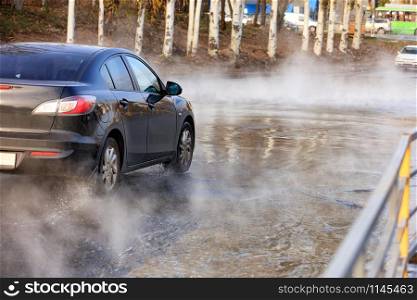 Accident on a heating main, a break in a pipe with hot water, a black car driving along a flooded road, water vapor forms a smokescreen over the asphalt, an image with copy space.. A black car is driving along a hot-water-filled road after a breakthrough in the city heating main.