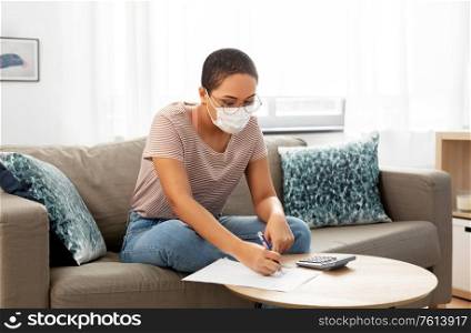accounting, taxes and finances concept - young african american woman in glasses with papers and calculator wearing face protective medical mask for protection from virus disease at home. woman in mask with papers and calculator at home