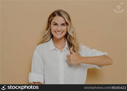 Achieving goals. Portrait of positive young woman with broad smile showing thumb up gesture with hand while posing in studio against beige wall, looking at camera with happy expression. Body language. Portrait of positive young woman with broad smile showing thumb up gesture with hand