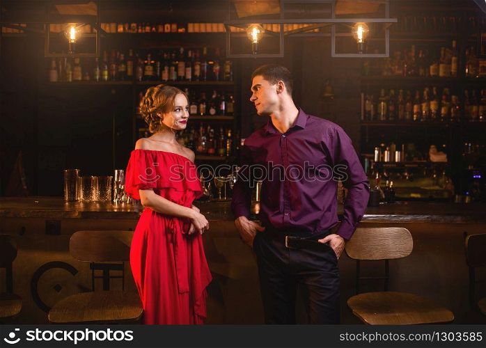 Acquaintance in bar, beautiful woman in red dress flirts with man behind counter. Date in nightclub, attractive couple funs together indoors. Acquaintance in bar, woman flirts with man