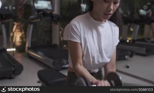 Active Asian woman in white top doing dumbbell curl while sitting on bench, real people exercising in fitness, fit girl controlling her own circles of breathing, fitness training, body condition