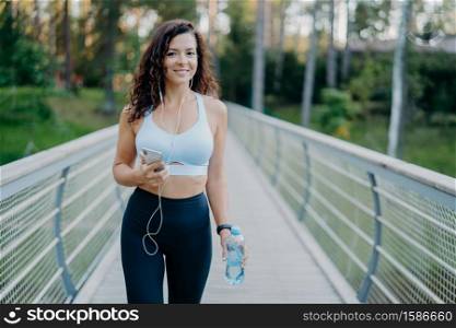 Active beautiful sporty woman in active wear holds mobile phone, listens music in earphones, takes break after jogging holds bottle of fresh water poses at bridge outdoors. Fitness, lifestyle concept