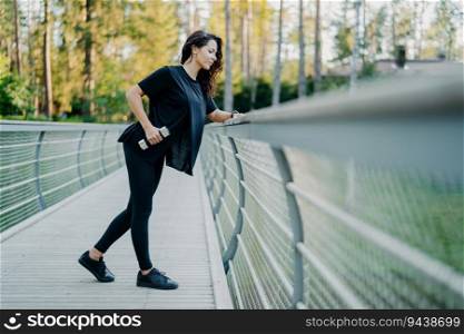 Active brunette in casual t-shirt and leggings holds dumbbells, poses at a bridge, goes for outdoor sports. Leading a healthy lifestyle, lifting weights, using sports equipment.