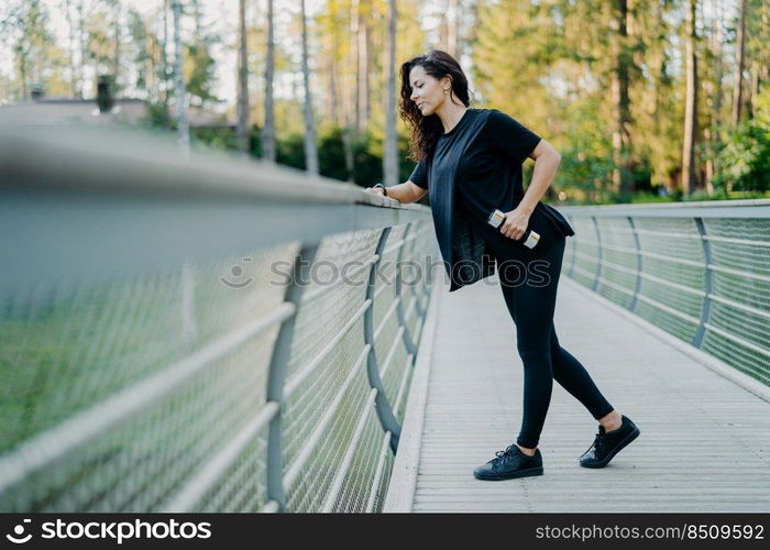 Active brunette woman dressed in casual t shirt and leggings, holds dumbbells, poses at bridge, looks down, goes in for sport outdoor, leads healthy lifestyle, lifts weight, uses sport equipment