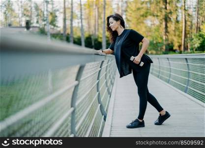Active brunette woman dressed in casual t shirt and leggings, holds dumbbells, poses at bridge, looks down, goes in for sport outdoor, leads healthy lifestyle, lifts weight, uses sport equipment