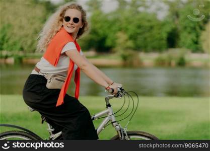 Active Caucasian curly woman looks aside, rides sport bicycle, covers distance and travels in open air, dressed in casual clothes, poses near small river with green trees, has healthy lifestyle