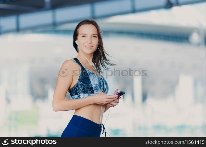Active Caucasian woman with dark hair, healthy skin, dressed in sportswear, uses up to date gadget, listens music with earphones, has charming smile, stands outdoor. People and lifestyle concept