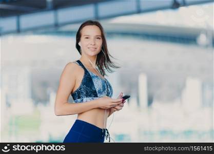 Active Caucasian woman with dark hair, healthy skin, dressed in sportswear, uses up to date gadget, listens music with earphones, has charming smile, stands outdoor. People and lifestyle concept