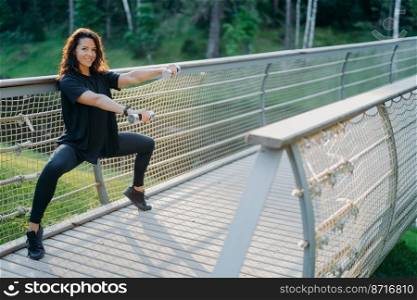 Active determined woman does squat exercise with dumbbells, poses in active wear at bridge, uses sport equipment, raises weight and trains biceps, has glad expression. Sporty lifestyle concept